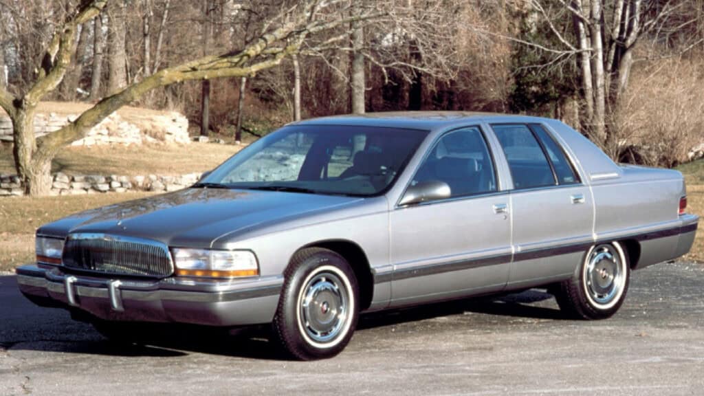 Silver vintage luxury sedan parked outdoors with trees and rocks in the background, showcasing classic car design elements.